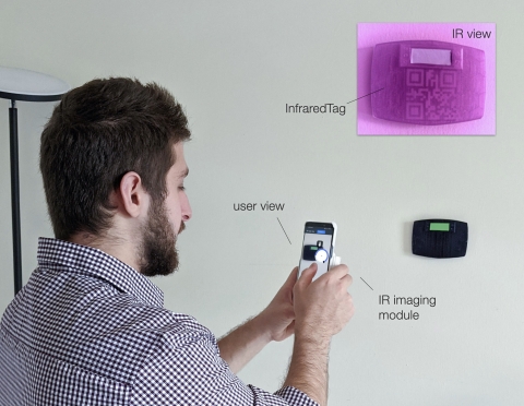A person holds a smartphone with an IR camera up to a thermostat mounted on a wall. An insert shows the infrared view of the thermostat with a previously invisible QR code visible 