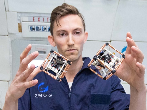 Two robotic cubes float in microgravity in front of a map in a blue jumpsuit